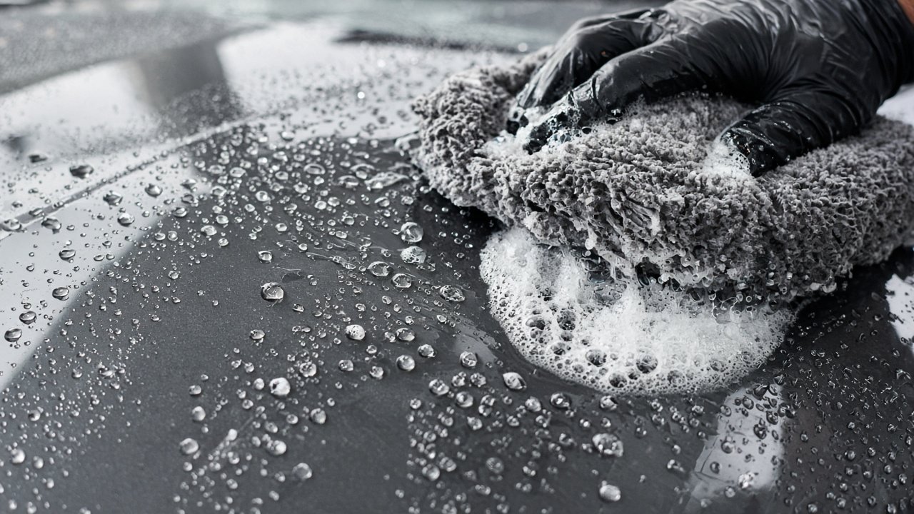 Gloved hand washing a car with a soft microfibre mitt and soapy foam