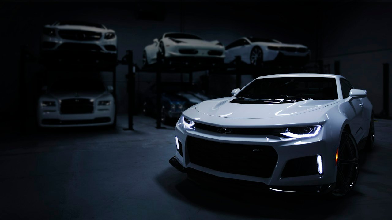 White sports car with its headlights on in a dim garage, with more cars on lifts behind it